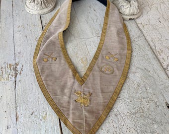 May include: A vintage, light tan ceremonial collar with gold trim and embroidered symbols. The collar has a black lining and a pointed bottom. The symbols include crossed tools and a letter "P" within a circle, and a gold emblem of a deer.