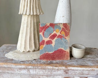 Può includere: Un piccolo blocco di legno quadrato con un motivo colorato di forme rosse, blu e gialle. Il blocco è esposto su una superficie di legno verniciata di bianco anticato, insieme a una piccola tazza di ceramica e due oggetti decorativi bianchi.