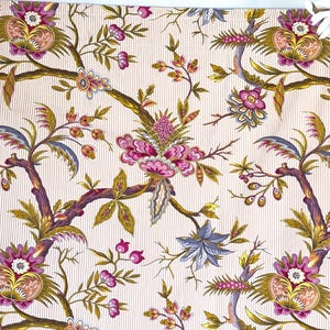 Op de afbeelding: Een witte stof met een herhalend patroon van roze, gele en blauwe bloemenontwerpen op een witte achtergrond met dunne, lichtblauwe strepen.