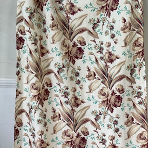 Könnte beinhalten: Ein weißer Stoff mit einem sich wiederholenden Muster aus braunen und grünen Blumendesigns. Der Stoff zeigt große, stilisierte Rosen und Blätter.