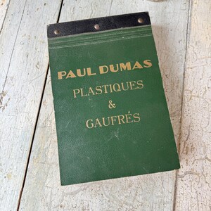 May include: A vintage green notepad with the text "PAUL DUMAS PLASTIQUES & GAUFRÉS" in gold lettering. The notepad has a black leather spine and is held together with metal rivets. The notepad is on a distressed, light-colored wooden surface.