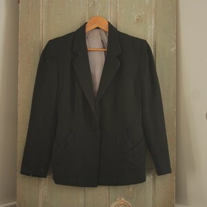 May include: A black blazer with a single button closure and a notched lapel. The blazer has two pockets on the front and is hanging on a wooden hanger.