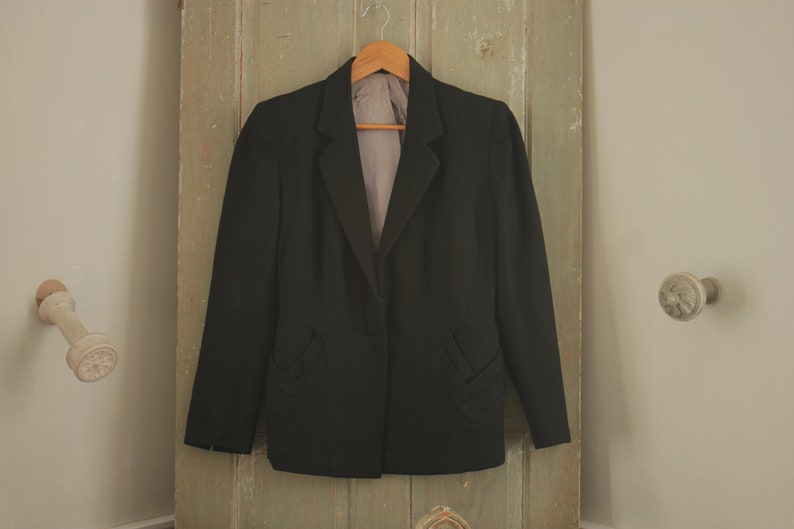 May include: A black blazer with a single button closure and a notched lapel. The blazer has two pockets on the front and is hanging on a wooden hanger.