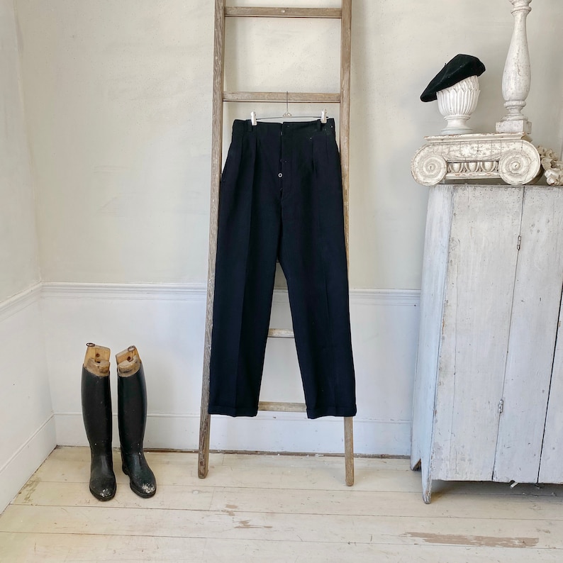 May include: Black high-waisted pants hanging on a wooden ladder. The pants have a single button closure and pleats at the front.