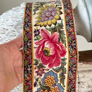 May include: A vintage floral trim with a cream background, featuring a pink flower, yellow flower, and blue and yellow floral design. The trim has a black and red border with a paisley pattern.
