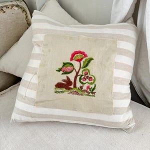 Può includere: Un cuscino a righe beige e bianco con un cuscino beige più piccolo all'interno. Il cuscino più piccolo ha un disegno ricamato di un coniglio seduto sotto un fiore rosa e verde.