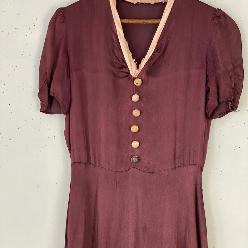 May include: A burgundy silk dress with a pink trim around the neckline and a row of buttons down the front.