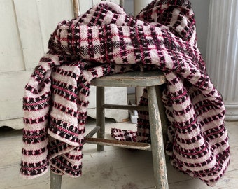 May include: A pink and black striped throw blanket draped over a wooden chair. The blanket has a woven texture and is made of a soft, cozy material.