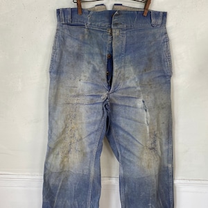 May include: Vintage blue denim work trousers with a button fly. The trousers show signs of wear and tear, including discolouration and holes. The trousers are hanging from a wooden clothes hanger.