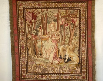 May include: A vintage tapestry with a woman sitting in a chair in a forest setting. The tapestry has a pink, green, and brown color scheme and features two dogs. The tapestry is framed with a wooden frame.