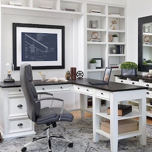 May include: White L-shaped desk with a dark wood top and two drawers on each side. A black office chair with a gray leather seat is positioned in front of the desk. A laptop computer is on the desk with a glass globe and a book on the shelf behind it.