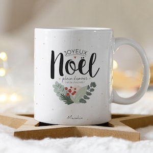 Op de afbeelding: Witte keramische mok met een feestelijk kerstontwerp. Het heeft de tekst "Joyeux Noël" in zwarte schrijfletters met een rood hart en de zin "plein d'amour (et de chocolat)" in kleinere tekst. De mok heeft een witte handgreep en een witte achtergrond met kleine witte stippen.