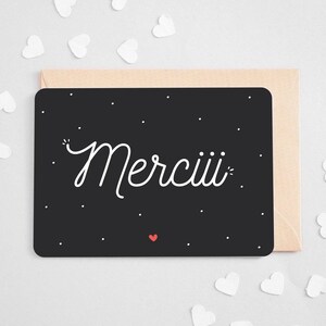May include: A black thank you card with white lettering that says 'Merciii' in a script font. The card has small white dots and a small red heart in the bottom left corner. The card is displayed on a light beige envelope and surrounded by small white paper hearts.
