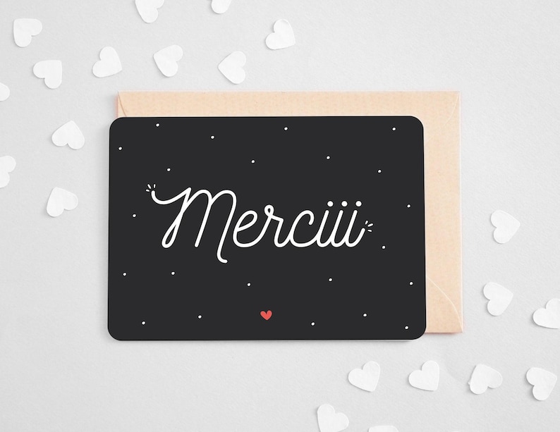 May include: A black thank you card with white lettering that says 'Merciii' in a script font. The card has small white dots and a small red heart in the bottom left corner. The card is displayed on a light beige envelope and surrounded by small white paper hearts.