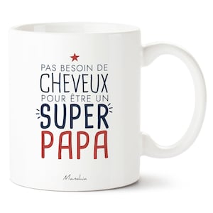 Peut inclure: Mug en céramique blanche avec une anse incurvée. Le mug affiche le texte "PAS BESOIN DE CHEVEUX POUR ÊTRE UN SUPER PAPA" en bleu et rouge, avec une étoile rouge au-dessus. La marque "Manahia" est en bas.
