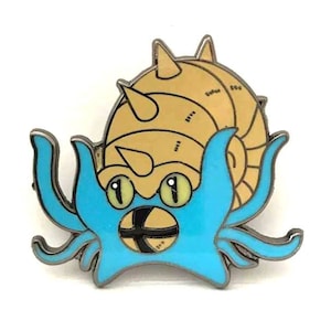 May include: A blue and gold enamel pin featuring a cartoon depiction of a Pokemon. The Pokemon has a blue body with a gold shell on its back and large, yellow eyes.