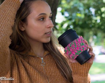 May include: A person wearing a brown sweater holds a black travel mug with a pink and green crocheted cozy. The cozy has the word "COZY" written on it.