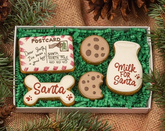 May include: A gift box filled with Christmas-themed cookies. The cookies include a postcard, a bone-shaped cookie with "Santa" written on it, two round cookies, and a milk bottle-shaped cookie with "Milk for Santa" written on it. The box is surrounded by pine needles.