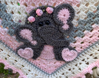 May include: A crocheted baby blanket in pink, white, and gray with a gray crocheted elephant applique. The elephant has pink ears and a pink heart on each foot. The elephant is decorated with pink flowers.