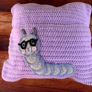 May include: A lavender crocheted pillow with a gray and white crocheted caterpillar wearing glasses. The caterpillar has a smiling face with pink cheeks.