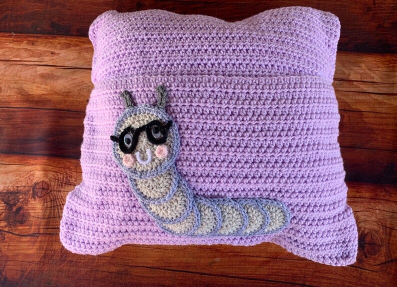 May include: A lavender crocheted pillow with a gray and white crocheted caterpillar wearing glasses. The caterpillar has a smiling face with pink cheeks.