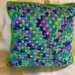 May include: A crocheted pillow with a green border and a colorful geometric pattern in shades of blue, green, purple, and white.