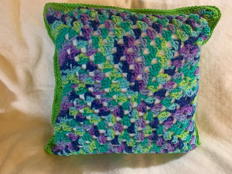 May include: A crocheted pillow with a green border and a colorful geometric pattern in shades of blue, green, purple, and white.