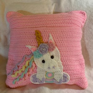 May include: A pink crocheted pillow with a white unicorn applique. The unicorn has a yellow horn, purple and blue mane, and pink and blue hooves.
