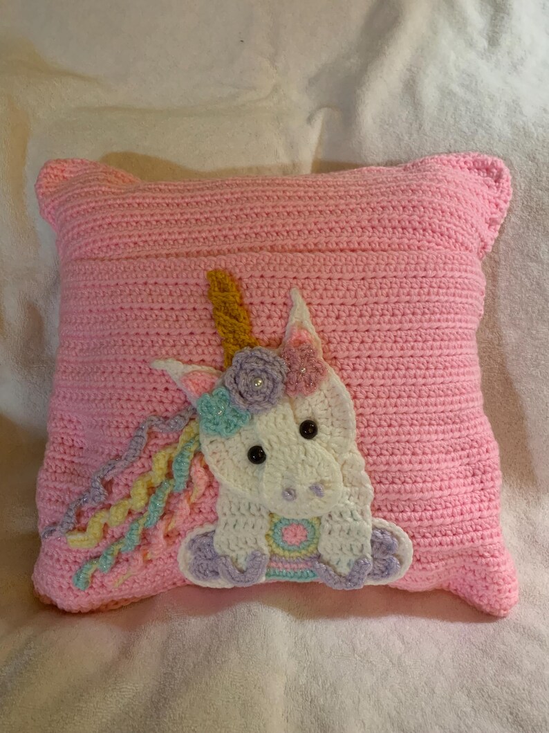 May include: A pink crocheted pillow with a white unicorn applique. The unicorn has a yellow horn, purple and blue mane, and pink and blue hooves.