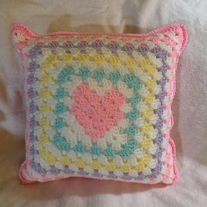 May include: A crocheted pillow with a heart design in the center. The pillow is made with pastel colors, including pink, yellow, green, and purple.