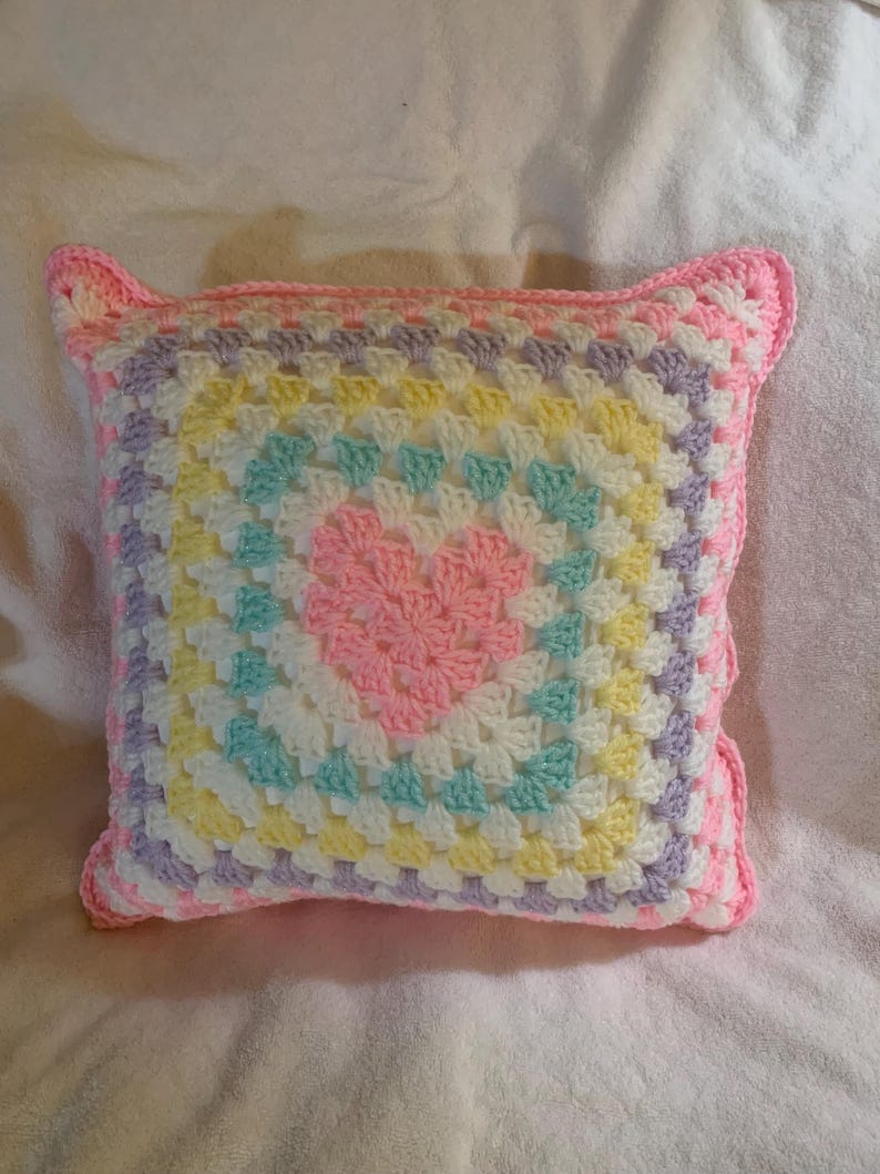 May include: A crocheted pillow with a heart design in the center. The pillow is made with pastel colors, including pink, yellow, green, and purple.