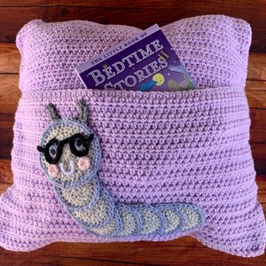 May include: A lavender crocheted pillow with a pocket for a book. The pillow has a gray crocheted caterpillar with glasses on the front. The book in the pocket is titled "The Little Book of Bedtime Stories".