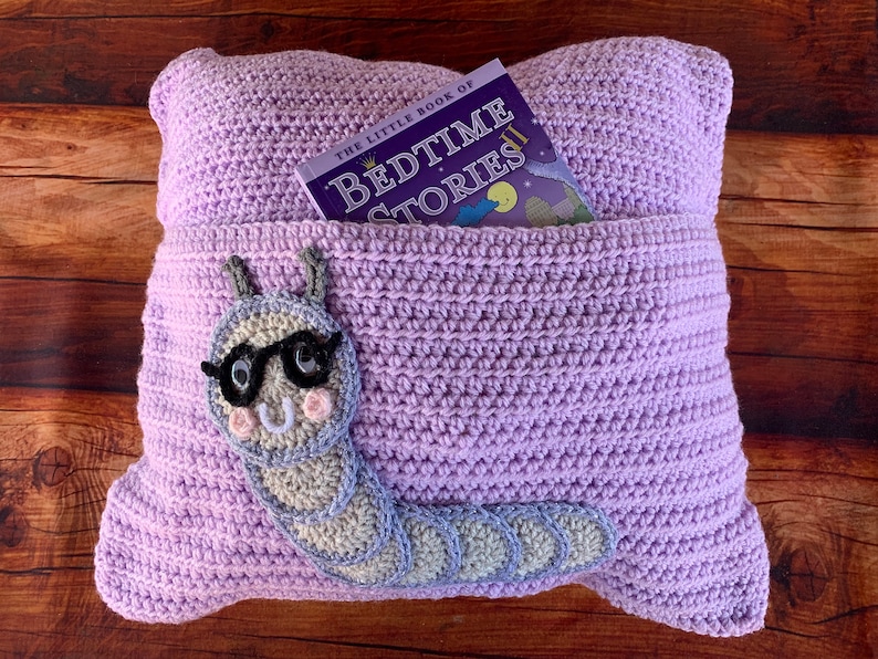 May include: A lavender crocheted pillow with a pocket for a book. The pillow has a gray crocheted caterpillar with glasses on the front. The book in the pocket is titled "The Little Book of Bedtime Stories".