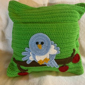 May include: A green crocheted pillow with a blue bird and red apples on a branch. The bird has white and yellow accents.