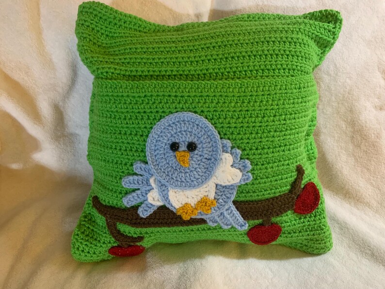 May include: A green crocheted pillow with a blue bird and red apples on a branch. The bird has white and yellow accents.
