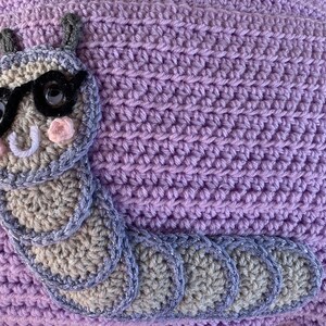 May include: A purple crocheted pillow with a gray crocheted caterpillar wearing sunglasses. The caterpillar has a white smile and pink cheeks.