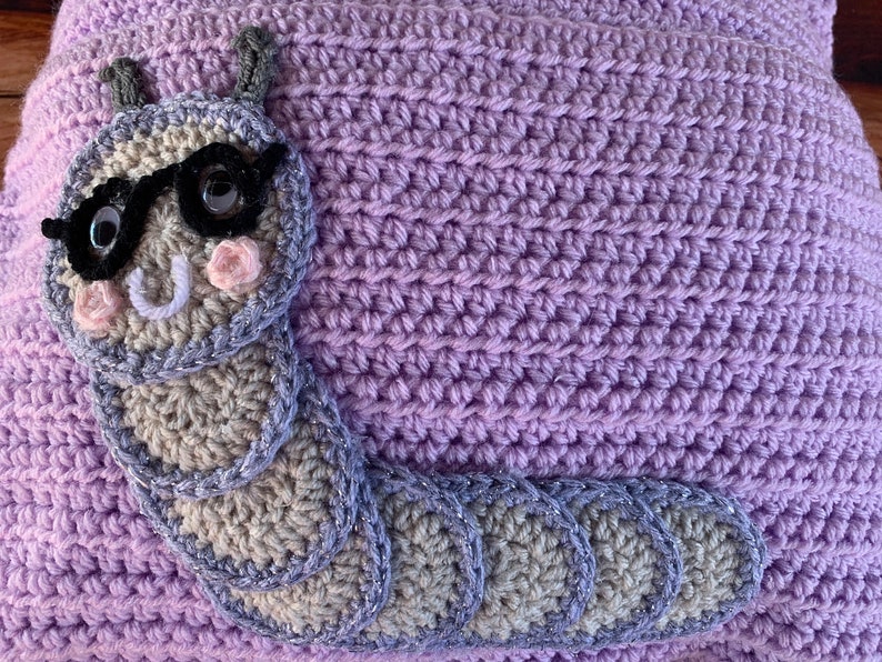 May include: A purple crocheted pillow with a gray crocheted caterpillar wearing sunglasses. The caterpillar has a white smile and pink cheeks.