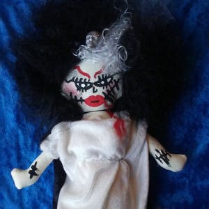 May include: A white fabric doll with black hair, a white dress, and red painted lips. The doll has black painted stitches on its face and arms. The doll is wearing red shoes.