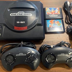 May include: A black Sega Genesis console with two controllers, two game cartridges, a power adapter, and AV cables. The console has the Sega Genesis logo and 16-BIT text. The game cartridges feature Sonic the Hedgehog.