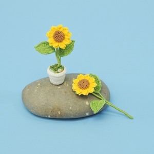 May include: Two crocheted sunflowers, one in a white pot, on a gray rock against a light blue background. The sunflowers are yellow with brown centers and green leaves.