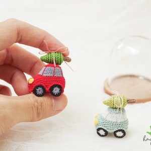 May include: Two miniature crochet cars, one red and one light blue, each with a green tree on top. The red car has a grey roof and black wheels. The light blue car has a white roof and black wheels. The cars are small enough to be held in a hand.
