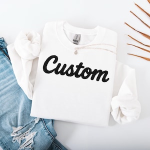 Peut inclure: Un sweat-shirt blanc avec le mot "Custom" en écriture noire. Le sweat-shirt est associé à un jean bleu clair déchiré et à un collier en argent. Une feuille de palmier séchée ajoute un élément naturel à la composition.
