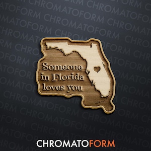 May include: Wooden plaque in the shape of Florida, featuring the text "Someone in Florida loves you." The state outline is light-coloured, with a heart marking a location. The plaque is made of wood and has a dark background.
