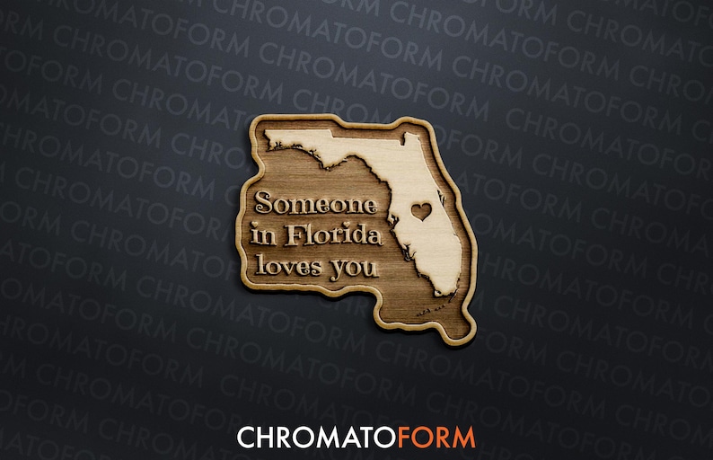 May include: Wooden plaque in the shape of Florida, featuring the text "Someone in Florida loves you." The state outline is light-coloured, with a heart marking a location. The plaque is made of wood and has a dark background.