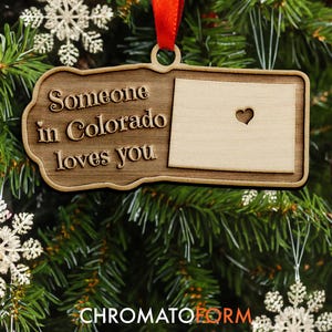 May include: Wooden Christmas ornament with the text "Someone in Colorado loves you." The ornament features the state of Colorado with a heart cutout. It hangs from a red ribbon on a Christmas tree with red and white baubles.
