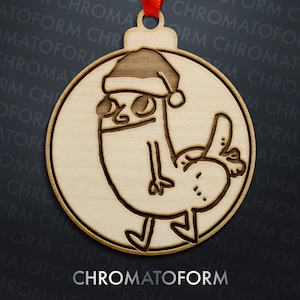 Pode incluir: Enfeite de Natal de madeira com um personagem de desenho animado usando um gorro de Papai Noel. O enfeite redondo tem uma fita vermelha para pendurar. A palavra "CHROMATOFORM" está impressa na parte inferior.