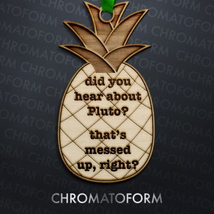 May include: Wooden pineapple-shaped ornament with the text "did you hear about Pluto? that's messed up, right?" and a green ribbon for hanging. The ornament is made of light brown wood and has a textured surface.