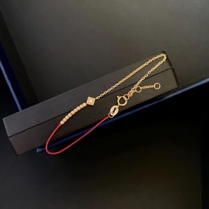 May include: Gold bracelet featuring a diamond-shaped charm with a small diamond, a series of gold beads, and a red cord. The bracelet has a delicate chain and clasp.