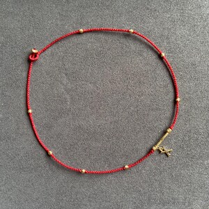 Può includere: Collana con cordino rosso con piccole perline dorate e un ciondolo dorato. La collana è annodata a un'estremità e ha una piccola barra dorata con un ciondolo all'altra estremità. La collana è esposta su una superficie grigia.