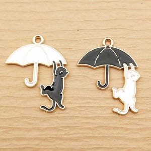 May include: Two gold-toned metal charms featuring a cat hanging from an umbrella. One charm has a black cat under a white umbrella, the other has a white cat under a black umbrella. The charms have a loop at the top for hanging.